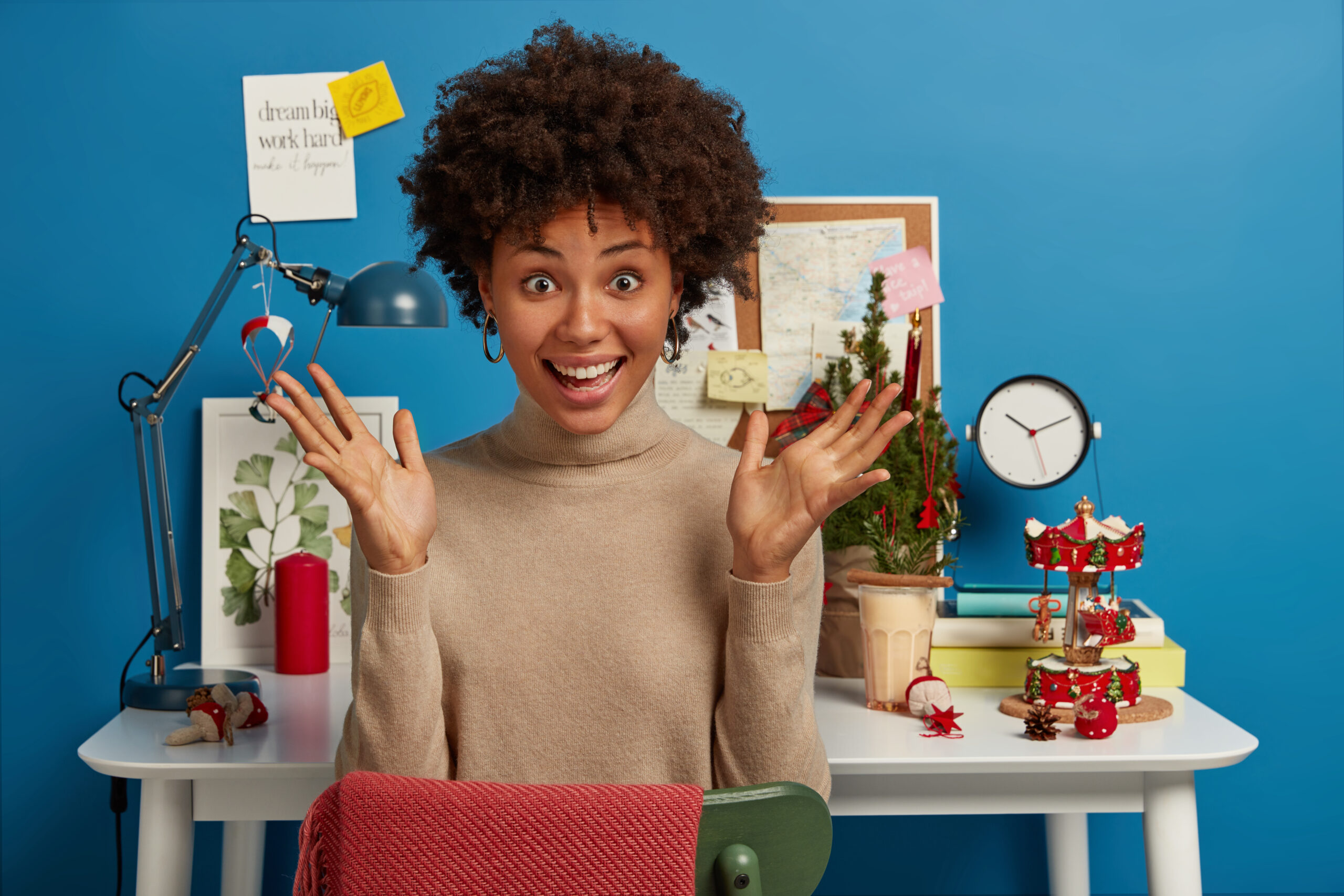 Yes, my task is completed! Joyful upbeat dark skinned woman has both palms raised, sits at desktop with Christmas tree and other holiday attributes for festive mood during work, gets good news