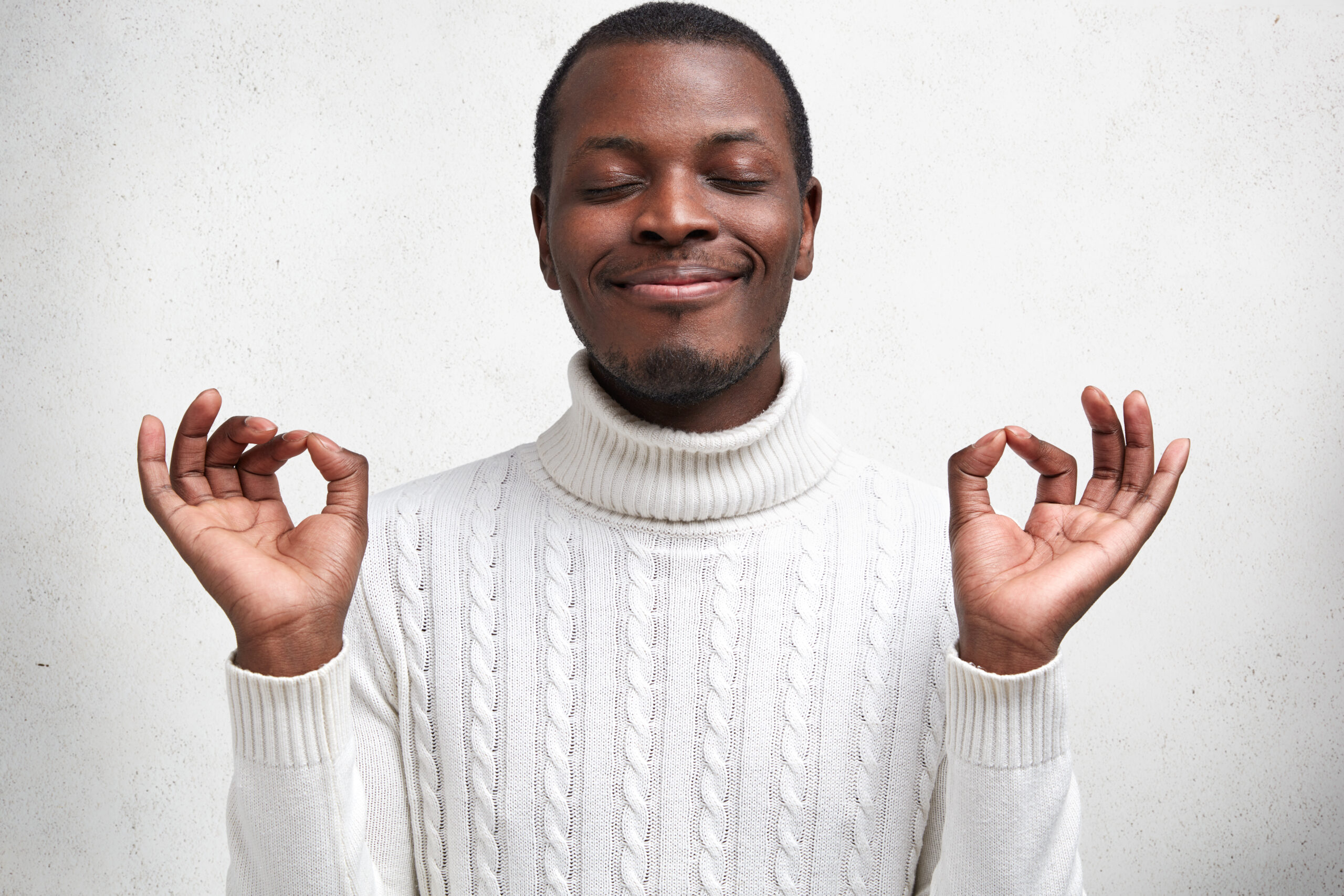 Content dark skinned young male keeps eyes shut and fingers in mudra sign, has calm expression, tries to relax after tired day, wears white sweater, poses against concrete studio background.