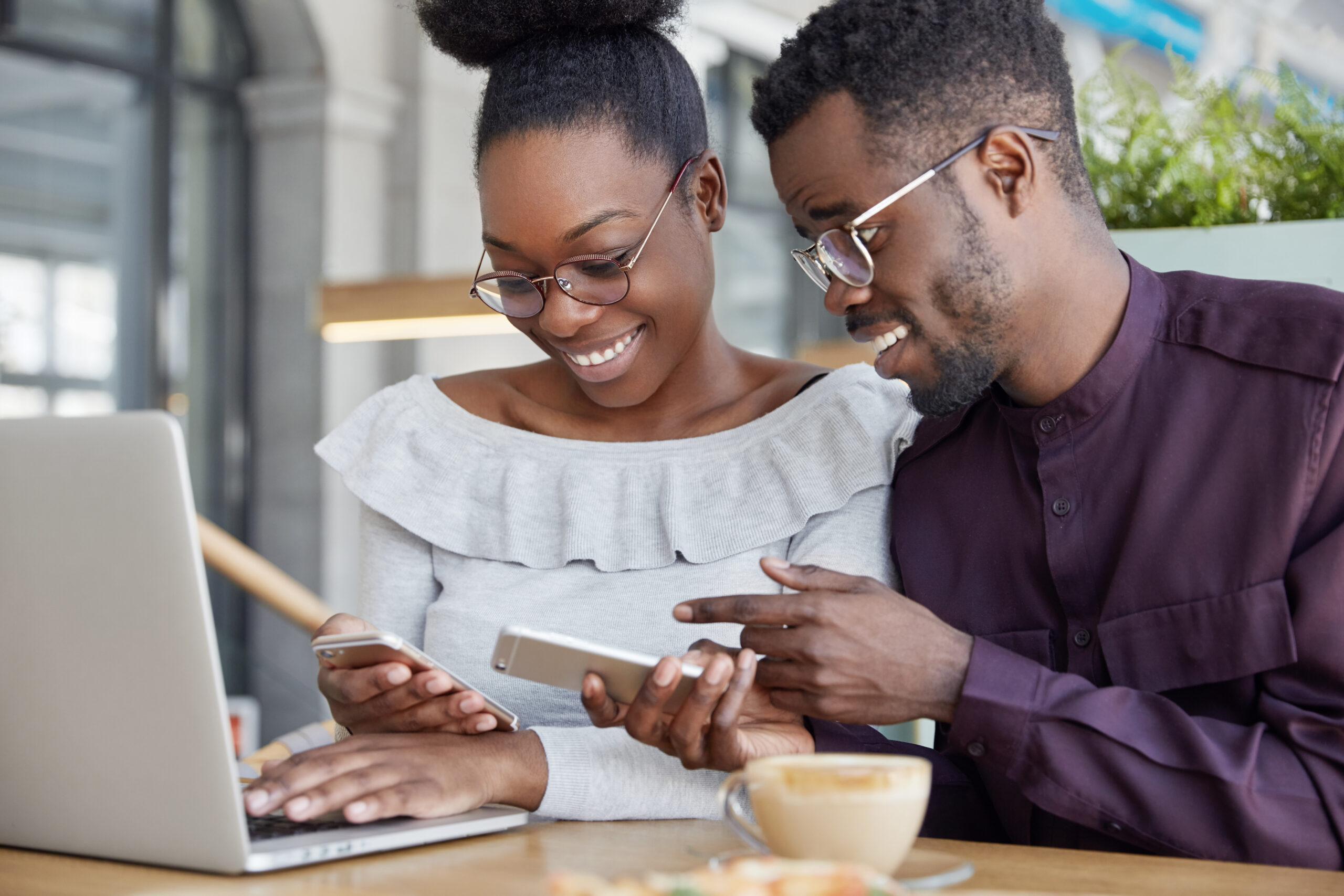 Black woman and man have informal meeting, glad to view photos on smart phone, wear spectacles, work together at common project via laptop computer, drink coffee. People and technology concept