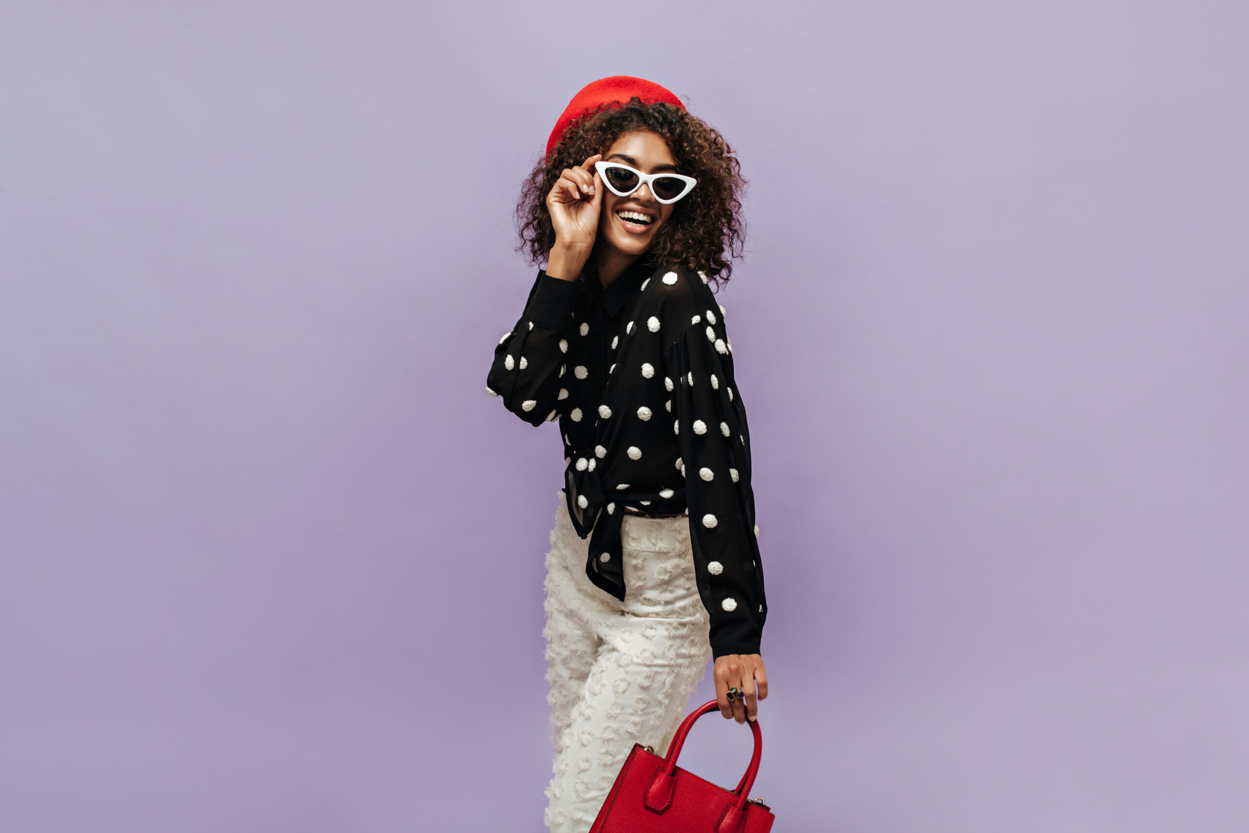Cheerful brunette haired woman with red cap and white sunglasses in black shirt and light pants smiling and holding handbag.