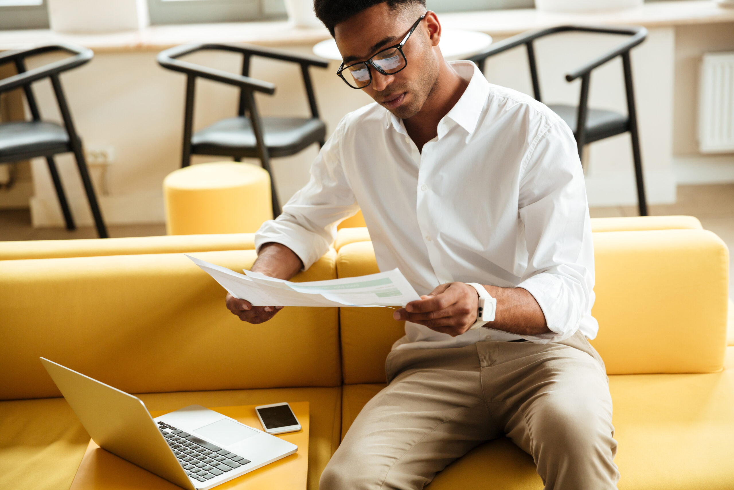Image of concentrated young african man sitting coworking by laptop computer. Looking aside holding documents.