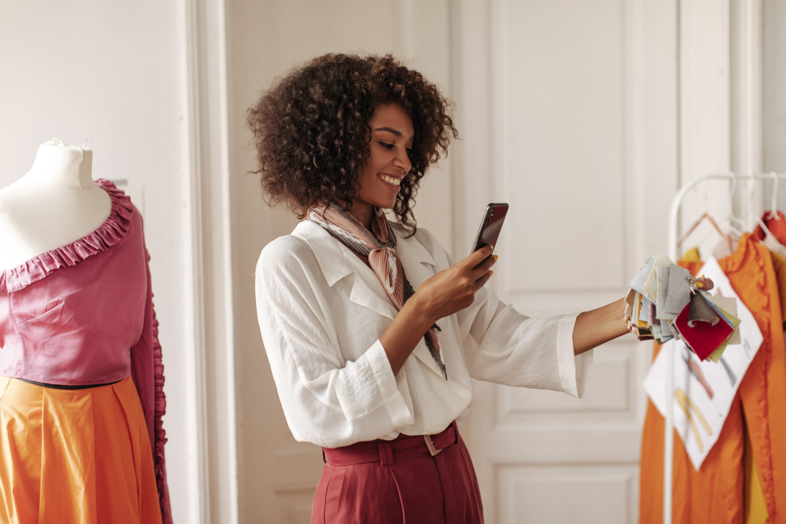 Happy brunette curly excited woman in white stylish blouse and burgundy pants holds phone, smiles and takes photo of textile samples.
